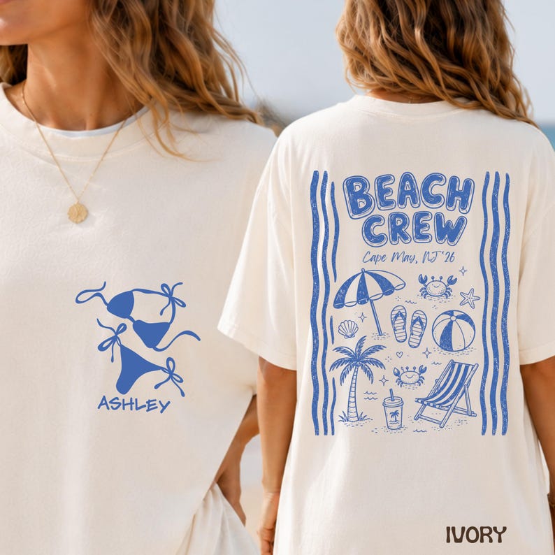 May include: An ivory t-shirt featuring blue graphics. The front displays a bikini and the name ASHLEY. The back shows the text BEACH CREW, Cape May, NJ '26, with illustrations of a beach umbrella, palm tree, flip-flops, ball, and chair.
