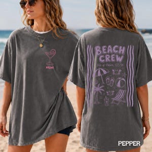 May include: A charcoal gray short-sleeve t-shirt featuring a pink martini glass graphic and the word MOM on the front. The back displays purple graphics including a beach umbrella, palm tree, beach chair, and the text BEACH CREW Isle of Palms, SC '26.
