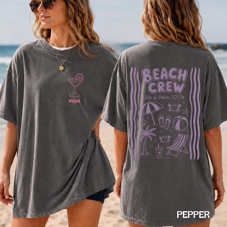 May include: A charcoal gray short-sleeve t-shirt featuring a pink martini glass graphic and the word MOM on the front. The back displays purple graphics including a beach umbrella, palm tree, beach chair, and the text BEACH CREW Isle of Palms, SC '26.