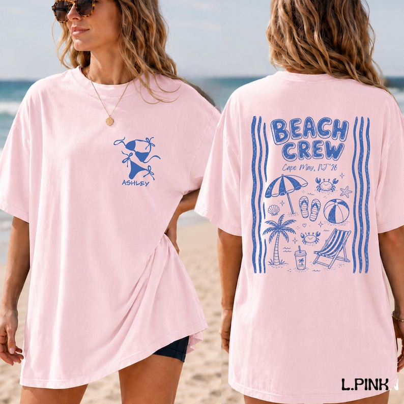 May include: A light pink t-shirt featuring a blue graphic of a bikini and the name Ashley on the front. The back displays a blue beach-themed design with the text BEACH CREW, Cape May, NJ '26, a beach umbrella, palm tree, and chair.