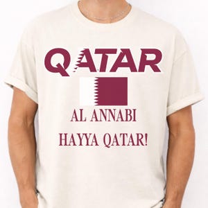 Ivory T-shirt showing a vibrant Qatar soccer flag graphic and Hayya Qatar chant print.