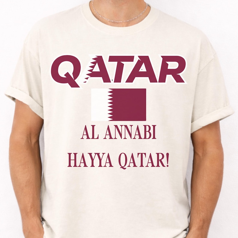Ivory T-shirt showing a vibrant Qatar soccer flag graphic and Hayya Qatar chant print.