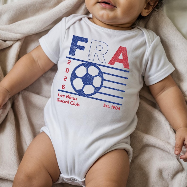 A flat-lay lifestyle shot of an infant wearing a soft white cotton baby bodysuit featuring the blue and red distressed retro French soccer club graphic, resting on a plush, wrinkled beige blanket.