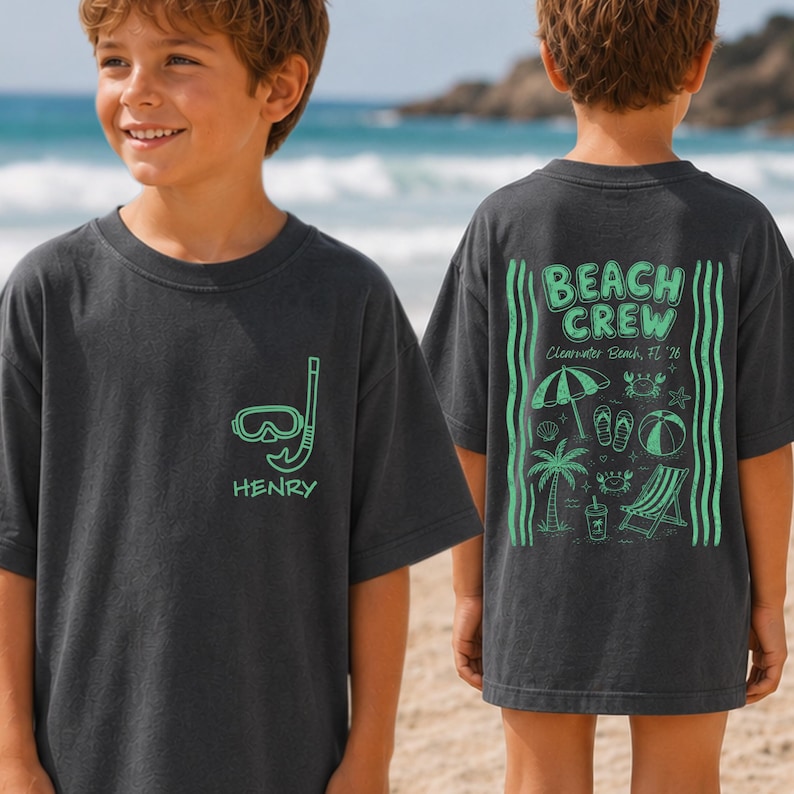 May include: A charcoal gray t-shirt featuring a light green snorkel mask and the name Henry on the front. The back displays a light green graphic with the text Beach Crew, Clearwater Beach, FL '26, including icons of a palm tree, umbrella, and chair.