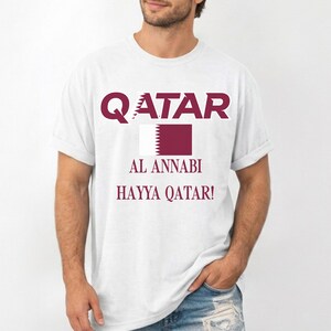 May include: White t-shirt with maroon text and a maroon and white flag design. The word "QATAR" is at the top, with "AL ANNABI" and "HAYYA QATAR!" below the flag. The shirt is a casual style.