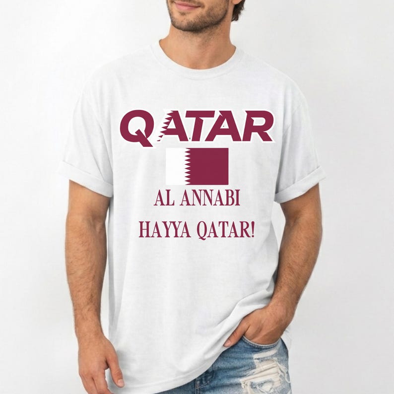 May include: White t-shirt with maroon text and a maroon and white flag design. The word "QATAR" is at the top, with "AL ANNABI" and "HAYYA QATAR!" below the flag. The shirt is a casual style.