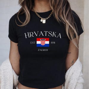 May include: Black t-shirt with the word "HRVATSKA" in white, above a Croatian flag graphic. Below the flag are the words "EST 1991 ZAGREB". The shirt has short sleeves and a crew neck.