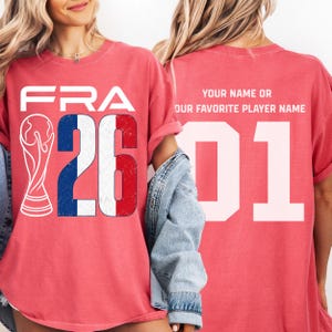 Puede incluir: Camiseta roja con "FRA" y el n&uacute;mero "26" en blanco, con la bandera francesa incorporada en el dise&ntilde;o. Un gr&aacute;fico blanco de un trofeo est&aacute; a la izquierda. La parte trasera de la camiseta tiene el n&uacute;mero "01" y texto.