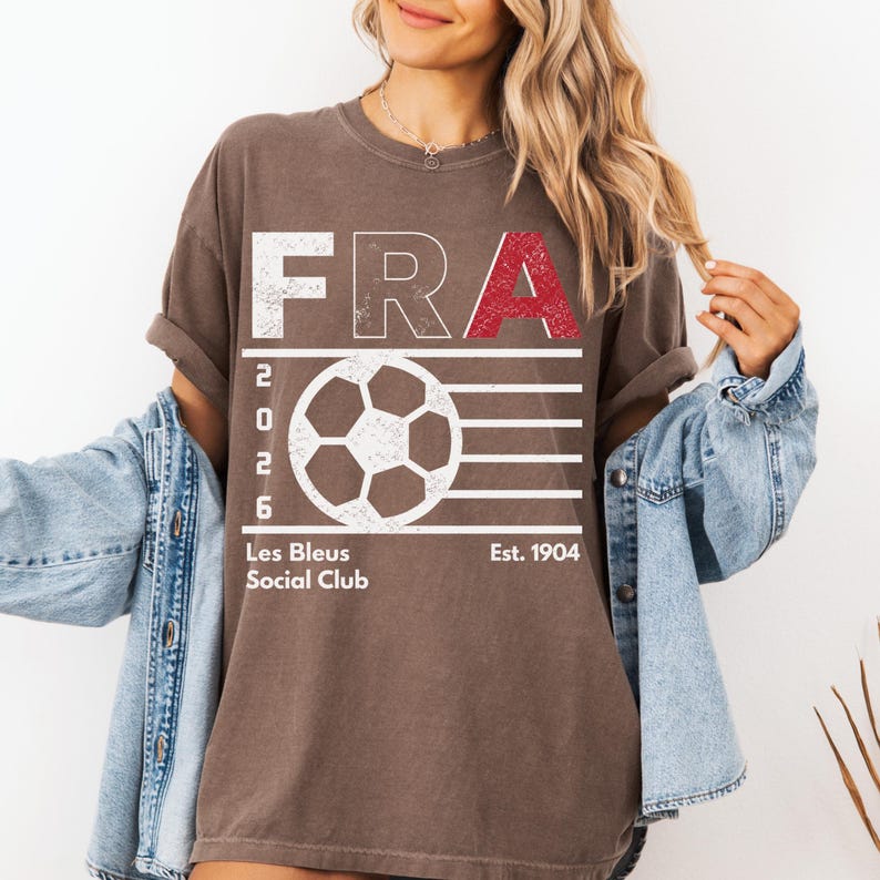 A female model styled in a casual streetwear look, wearing a vintage-ESPRESSO t-shirt with the white and red "FRA" soccer graphic, layered with an oversized denim jacket off her shoulders.