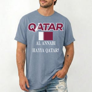 May include: A light blue t-shirt with the word "QATAR" in maroon letters above a maroon and white flag graphic. Below the flag are the words "AL ANNABI" and "HAYYA QATAR!" in white.