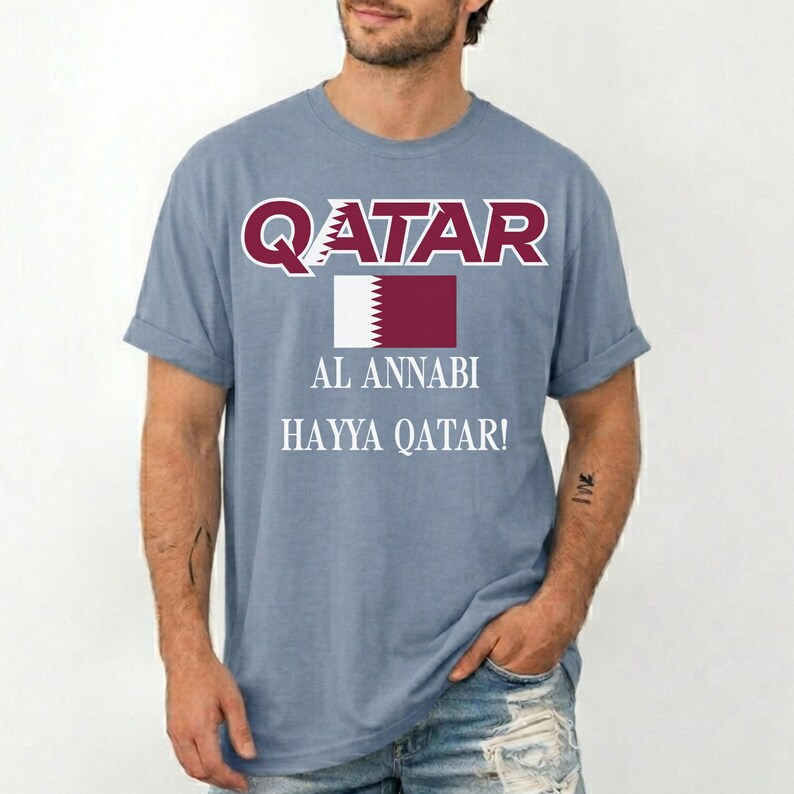 May include: A light blue t-shirt with the word "QATAR" in maroon letters above a maroon and white flag graphic. Below the flag are the words "AL ANNABI" and "HAYYA QATAR!" in white.