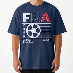 Peut inclure: Un t-shirt bleu marine avec un motif blanc et rouge &agrave; l'aspect vieilli. Le design comprend le texte FRA, l'ann&eacute;e 2026, un ballon de football, ainsi que les inscriptions Les Bleus Social Club et Est. 1904. Le graphisme pr&eacute;sente une texture us&eacute;e.