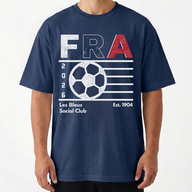 Peut inclure: Un t-shirt bleu marine avec un motif blanc et rouge &agrave; l'aspect vieilli. Le design comprend le texte FRA, l'ann&eacute;e 2026, un ballon de football, ainsi que les inscriptions Les Bleus Social Club et Est. 1904. Le graphisme pr&eacute;sente une texture us&eacute;e.