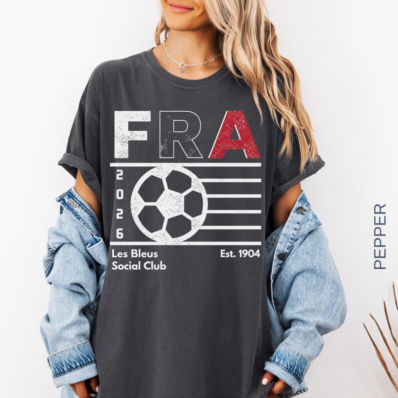 A front-facing lifestyle shot of a model wearing a dark charcoal gray mineral-washed t-shirt displaying the distressed white and red France 2026 soccer layout, styled with a light-blue denim jacket.
