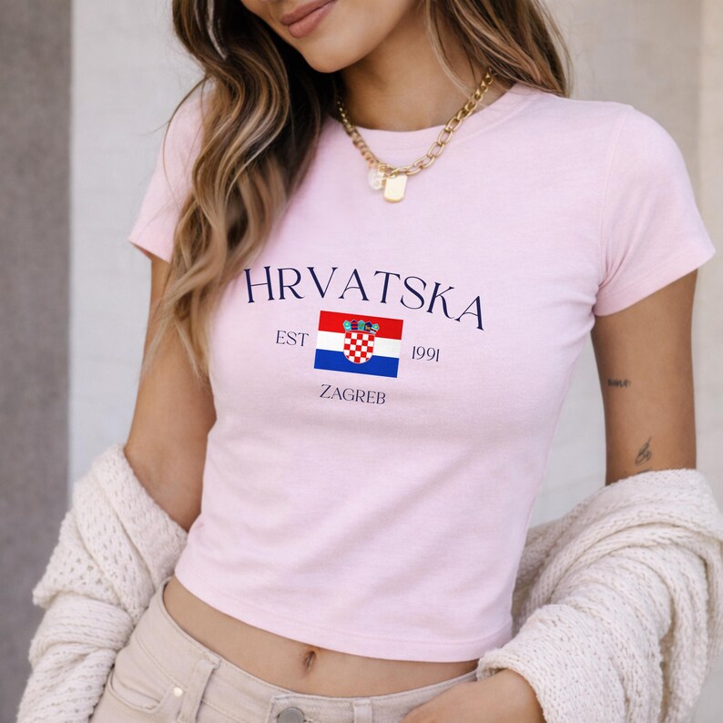 Close-up of a blonde woman wearing a fitted pink crop top with a Croatian flag and text reading Hrvatska EST 1991 Zagreb. She wears a chunky gold chain necklace and a cozy cream knit cardigan draped off her shoulders.