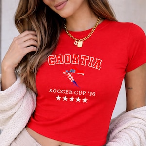 Close-up of a model wearing a red Croatia baby tee with a gold chain and cream knit cardigan. The collegiate "CROATIA" text and flag map are centered in soft daylight.