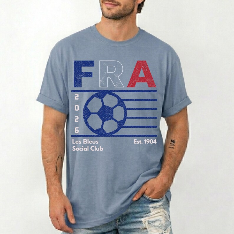 Peut inclure: Un t-shirt bleu d&eacute;lav&eacute; &agrave; manches courtes avec un motif graphique comprenant le texte FRA, un ballon de football, l'ann&eacute;e 2026 et les mots Les Bleus Social Club Est. 1904. Le design pr&eacute;sente une texture us&eacute;e en bleu, blanc et rouge.