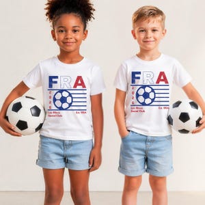 Two young children, a girl with curly hair and a boy, standing side-by-side holding classic black-and-white soccer balls, both wearing matching white youth t-shirts with the vintage blue and red France football graphic.