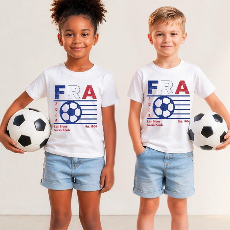 Two young children, a girl with curly hair and a boy, standing side-by-side holding classic black-and-white soccer balls, both wearing matching white youth t-shirts with the vintage blue and red France football graphic.