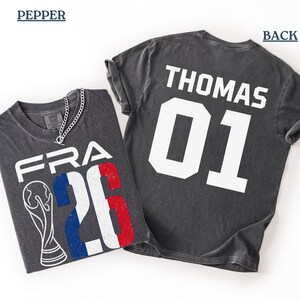 High-angle flat-lay product presentation showcasing a Comfort Colors Pepper grey t-shirt against a clean white background. The folded shirt on the left shows the distressed front design with a silver chain necklace styled over the neckline. The flat-spread shirt on the right exhibits the personalized back preview reading "THOMAS 01" in contrasting white athletic text.
