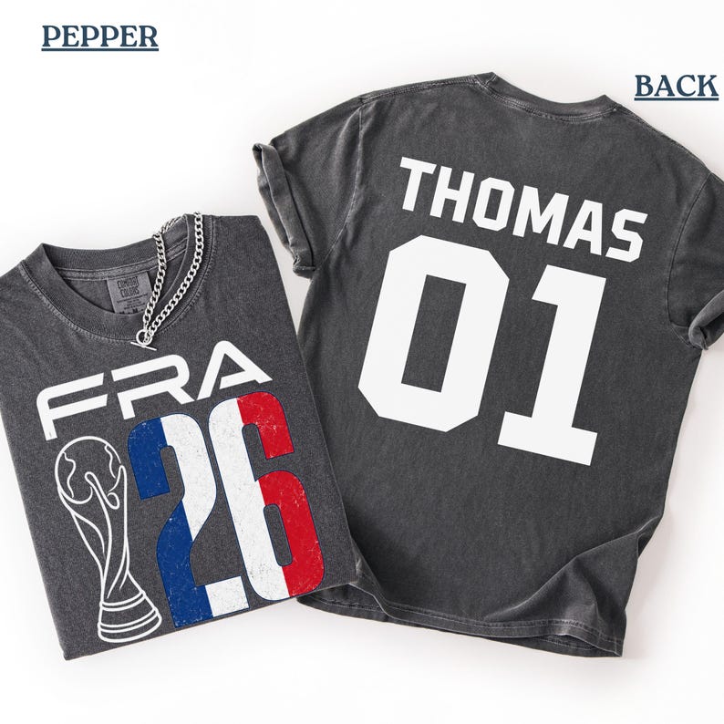 High-angle flat-lay product presentation showcasing a Comfort Colors Pepper grey t-shirt against a clean white background. The folded shirt on the left shows the distressed front design with a silver chain necklace styled over the neckline. The flat-spread shirt on the right exhibits the personalized back preview reading "THOMAS 01" in contrasting white athletic text.