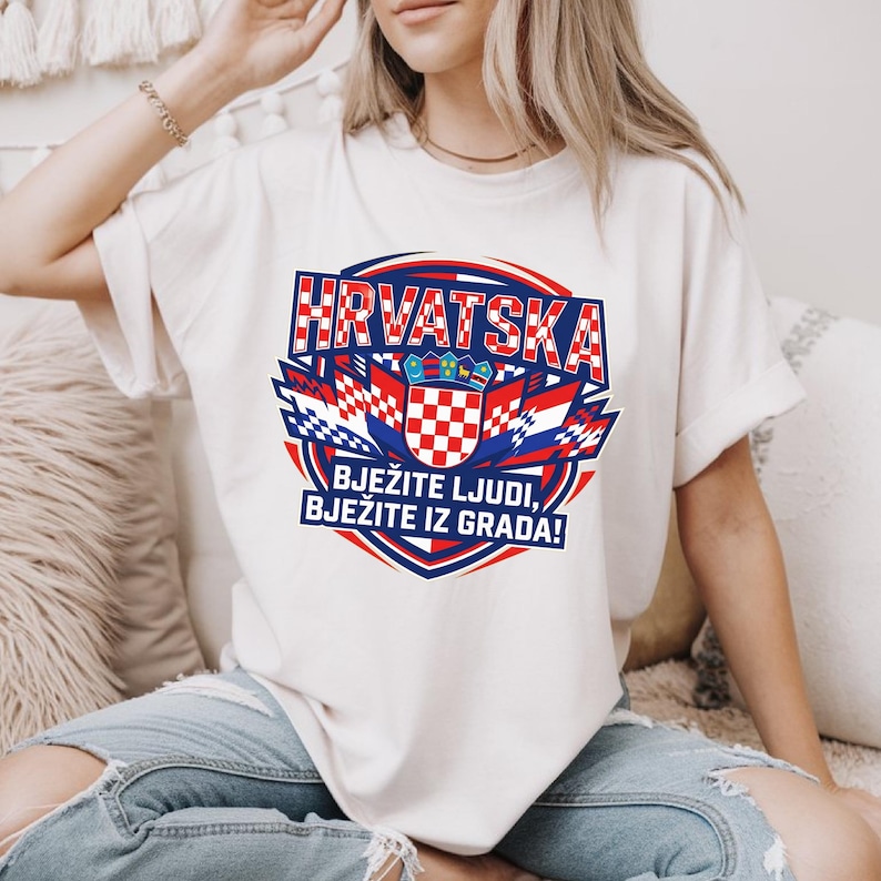 Oversized ivory cream t-shirt with a vibrant red and white chequy Croatia football crest graphic design, worn by a blonde woman sitting in a cozy bohemian room.