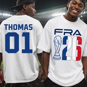 Front and back split preview featuring two male models in a brightly lit stadium environment. The model on the right smiles while facing forward, displaying the white graphic t-shirt with a distressed French flag texture inside the "26" block numbers. The model on the left is turned back, showing the personalized text layout "THOMAS 01" printed cleanly across the shoulders.