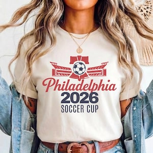 May include: Beige t-shirt with a red and blue graphic design. The design features a soccer ball, stadium, and the words "Philadelphia 2026 Soccer Cup" in a stylized font. The shirt has short sleeves.