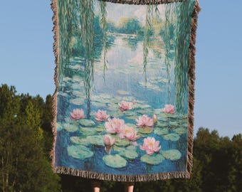 May include: A woven throw blanket depicting a water lily pond scene with pink flowers, green lily pads, and weeping willow trees. The blanket has a blue and green colour palette with a fringed edge. The blanket is held up against a blue sky.