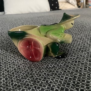 May include: A ceramic planter in the shape of a person pushing a wheelbarrow. The figure is wearing a green hat and jacket, with a light green and beige wheelbarrow. The wheel has a red center. The planter is on a gray patterned surface.