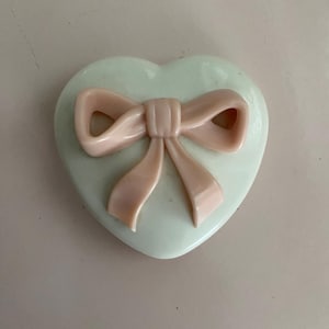 May include: A heart-shaped trinket box with a pale green base and a pink bow. The bow is centered on the heart, adding a decorative touch. The box is likely used for storing small items.