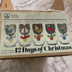 May include: A cardboard box for Anchor Hocking 12 Days of Christmas beverage glasses. The box features illustrations of five glasses, each depicting a scene from the song with colorful trees, birds, and banners labeled 1st Day through 5th Day.