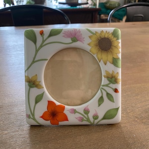 May include: A square white ceramic picture frame featuring a circular center opening. The frame is decorated with a colorful botanical pattern of yellow sunflowers, a bright orange flower, green leaves, and small pink flower buds on thin stems.