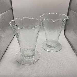 May include: Two clear glass dessert dishes with a textured surface and a scalloped rim. Each dish has a circular base and a flared, cone-shaped bowl. The dishes are empty and sit on a white surface.