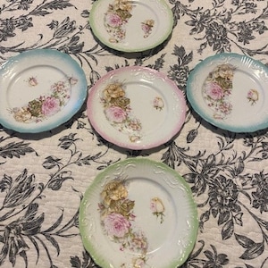 May include: Set of four vintage floral patterned plates with green, blue, and pink trim. The plates feature a delicate floral design with pink and white flowers.