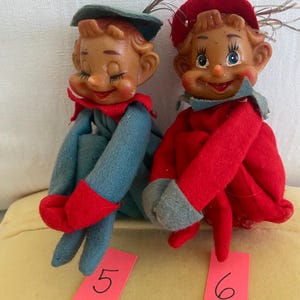 May include: Two elf dolls with molded plastic faces and felt clothing. The left doll wears a blue outfit with a green hat and red collar, labeled with a pink tag numbered 5. The right doll wears a red outfit with a red hat and blue collar, labeled with a pink tag numbered 6.