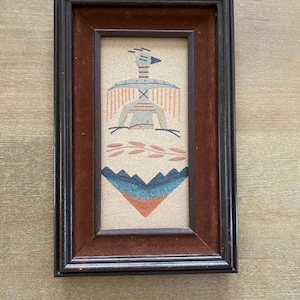 May include: A rectangular sand art piece in a dark brown wooden frame with a velvet border. The artwork features a stylized bird figure in blue, orange, and white, positioned above a leafy branch and a mountain range depicted in blue and orange tones.