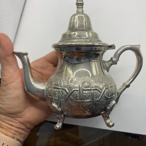 May include: A silver-colored metal teapot featuring an intricate embossed geometric and floral pattern on its rounded body. The teapot has a curved spout, a decorative lid with a finial, a handle, and stands on three small, ornate metal feet.