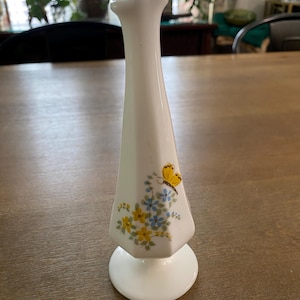 May include: A tall, white milk glass bud vase with a flared, ruffled rim and a circular base. The body of the vase features a painted floral design with small yellow and blue flowers and a single yellow butterfly perched near the top of the stems.