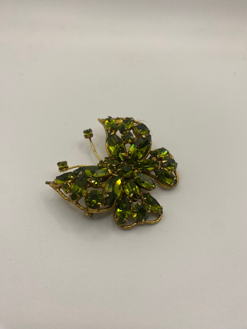 May include: A gold-toned butterfly brooch with green rhinestones. The brooch is made of metal and has a detailed design.