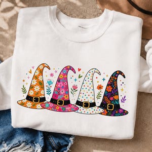 Könnte beinhalten: Ein weißes Sweatshirt mit Rundhalsausschnitt und einem Grafikdruck von vier nebeneinander angeordneten Hexenhüten. Jeder Hut hat ein eigenes Muster, darunter orangefarbene Gänseblümchen, rosa Blumen, bunte Punkte und ein dunkelviolettes Blumenmuster.