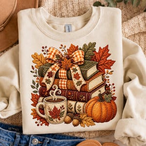 May include: A cream-colored crewneck sweatshirt featuring a graphic print of a stack of books tied with a plaid ribbon, a steaming mug decorated with a maple leaf, a pumpkin, and autumn foliage including orange, red, and brown leaves.