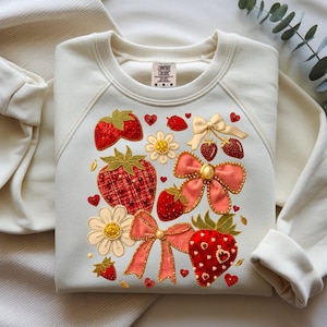 Op de afbeelding: Een crèmekleurig sweatshirt met ronde hals en een geborduurd ontwerp van rode aardbeien, roze en crèmekleurige strikken, witte bloemen en kleine rode hartjes. Het Comfort Colors-merklabel is zichtbaar bij de binnenhalslijn.