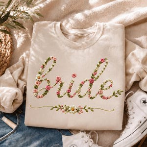 May include: A beige crewneck sweatshirt featuring the word Bride embroidered in a cursive script. The lettering is composed of small pink, white, and green floral motifs. A horizontal floral vine accent is positioned below the text.