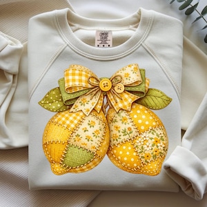 Puede incluir: Sudadera de color crema con un diseño de limón. Los limones son amarillos con patrones de patchwork, acentuados por un lazo y hojas a cuadros verdes y amarillos. El diseño incluye dos pequeños botones. La sudadera tiene la etiqueta de la marca Comfort Colors.