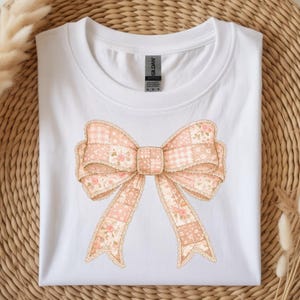 Peut inclure: Un t-shirt blanc à manches courtes orné d'un grand nœud imprimé au centre. Le nœud présente un motif patchwork dans des tons rose clair, beige et crème, combinant des imprimés floraux et des carreaux vichy avec une bordure en dentelle.