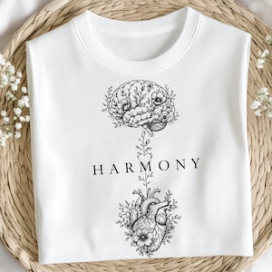 Può includere: Felpa bianca girocollo con grafica in bianco e nero. Il design presenta un cervello e un cuore collegati da uno stelo floreale, con la parola "HARMONY" al centro. Il cervello e il cuore sono circondati da elementi floreali.