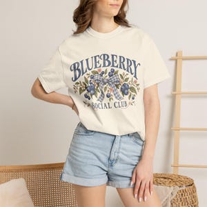 May include: A cream-coloured short-sleeve t-shirt featuring a graphic print of blueberries, green leaves, and a blue and white checkered bow. The text reads BLUEBERRY SOCIAL CLUB in a dark blue arched font above and below the botanical illustration.