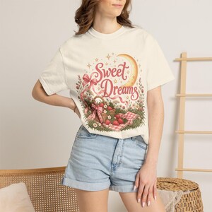 以下が含まれることがあります： クリーム色のTシャツ。赤いイチゴが入った籐のバスケット、ピンクのリボン、三日月、そして「Sweet Dreams」という文字が、小さな花や星と共にチェック柄のピクニックシートの上に描かれたグラフィックプリントが施されています。