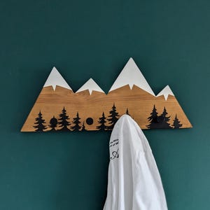 Puede incluir: Perchero de pared de madera con forma de montañas, con picos blancos y diseños de árboles negros. Una camiseta blanca cuelga de los ganchos. La pared está pintada de color verde azulado. El perchero mide unos 40 cm de ancho.